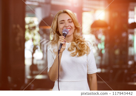Mature pretty blond woman singing in microphone soulfully indoors. Abstract blurred indoor background. 117943442