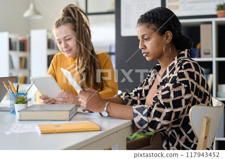 Side view portrait of ethnic female student watching online lesson on digital tablet while sitting at desk and studying during class in school or college 117943452