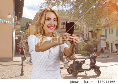 Portrait of happy blonde woman taking selfie on smartphone frontal camera outdoors. European street in the background. 117943466