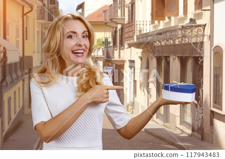 Happy blonde woman holding a blue heart shape gift box and pointing with index finger. European street in the background. Happy blonde woman holding a blue heart shape gift box and pointing with index finger. European street in the background. 117943483