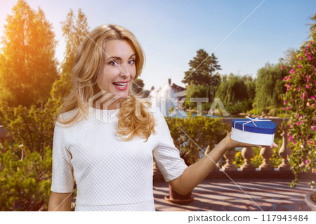 Happy caucasian blonde woman holding a blue heart shape gift box on her left palm. Summer park in the background. Happy caucasian blonde woman holding a blue heart shape gift box on her left palm. Summer park in the background. 117943484