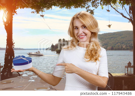 Portrait of mature blonde woman pointing with finger at the blue heart shape gift box. Nature landscape in the background. Portrait of mature blonde woman pointing with finger at the blue heart shape gift box. Nature landscape in the background. 117943486
