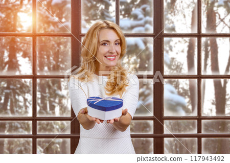 Portrait of blonde woman gives you a christmas present. Indoor window in the background with winter evening scape view. Portrait of blonde woman gives you a christmas present. Indoor window in the background with winter evening scape view. 117943492