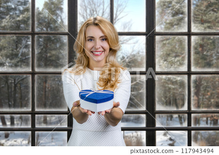 Blonde woman holding gives you a blue heart shape gift box. Indoor window in the background with winter scape view. 117943494