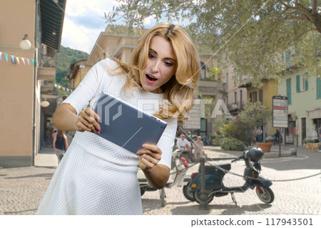 Excited mature blond woman standing outdoors playing a video game on the tablet device. Urban cityscape in the background. Excited mature blond woman standing outdoors playing a video game on the tablet device. Urban cityscape in the background. 117943501