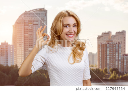 Portrait of a happy mature blond woman shows ok gesture sign. Residential area in the background. Portrait of a happy mature blond woman shows ok gesture sign. Residential area in the background. 117943534