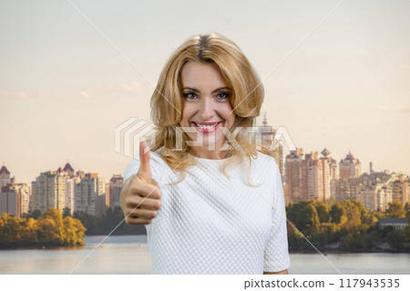 Portrait of a happy mature blond woman shows like gesture sign. Urban cityscape in the background. 117943535