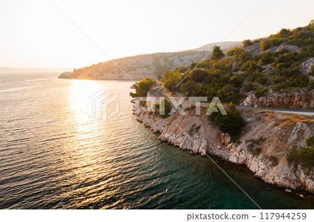 Sunset view of a winding coastal road alongside clear waters in a serene natural landscape. Sunset view of a winding coastal road alongside clear waters in a serene natural landscape. 117944259