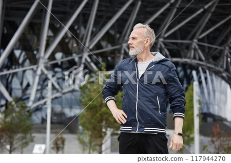 Medium shot of fit senior man in navy windbreaker standing with hand on waist resting after daily jogging workout at city park against steel stadium structures, copy space 117944270