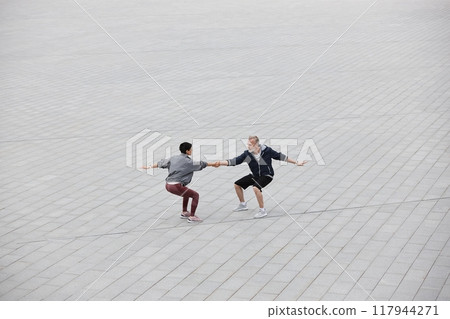 Wide angle shot of fit senior man and woman doing partner exercise balancing in seated position holding hands in middle of large tiled gray square at city park, copy space 117944271