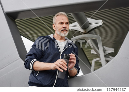 Medium shot of senior man with gray hair holding plastic bottle of water resting after daily jogging workout under shade pavilion at city park, copy space 117944287