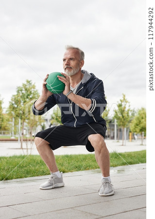 Vertical shot of fit senior man holding ball in static squat position working out at city park against gray autumn sky, copy space 117944292