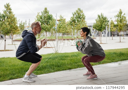 Side view of senior smiling woman and man in sportswear doing squat jumps passing ball Side view of senior smiling woman and man in sportswear doing squat jumps passing ball 117944295