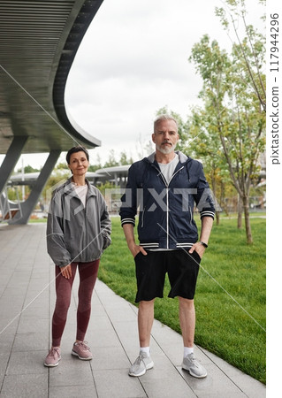 Vertical shot of senior smiling woman and man in sportswear on regular morning workout looking at camera and posing at city park, copy space Vertical shot of senior smiling woman and man in sportswear on regular morning workout looking at camera and posing at city park, copy space 117944296