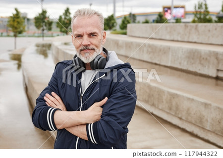 Medium portrait shot of smiling senior man in navy windbreaker on morning workout at city park posing with folded arms and looking at camera, copy space Medium portrait shot of smiling senior man in navy windbreaker on morning workout at city park posing with folded arms and looking at camera, copy space 117944322