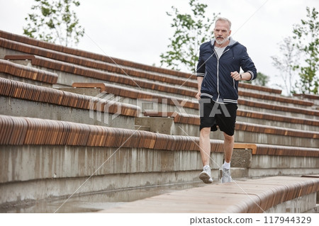 Full shot of fit elderly man in sportswear jogging along wooden benches of amphitheater working out regularly at city park, copy space Full shot of fit elderly man in sportswear jogging along wooden benches of amphitheater working out regularly at city park, copy space 117944329