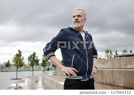 Chest up portrait of fit elderly man in sportswear with hands on waist looking away while resting after everyday jogging in city park against cloudy sky, copy space 117944333