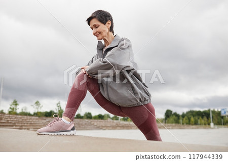Smiling fit elderly woman in pink leggings stretching leg muscles in lunge position against cloudy sky in city park, copy space 117944339