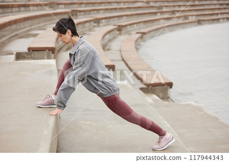 Side view of fit senior woman in pink sportswear stretching leg muscles on huge concrete steps of amphitheater getting ready to run jogging outside in city park, copy space 117944343