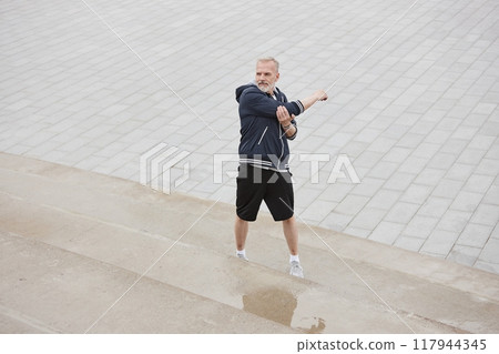 Wide angle shot of fit elderly man in sportswear stretching arms standing on concrete steps while getting ready to run jogging against background of tiled square in city park, copy space Wide angle shot of fit elderly man in sportswear stretching arms standing on concrete steps while getting ready to run jogging against background of tiled square in city park, copy space 117944345