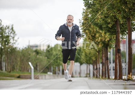 Full shot of fit elderly man in sportswear jogging along lane of trees working out regularly in morning city park, copy space 117944397