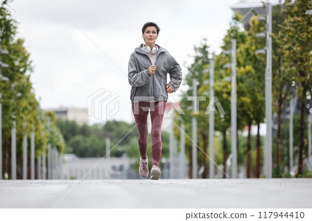 Full shot of fit elderly woman in sportswear jogging along wide walkway in morning city park working out on regular basis, copy space 117944410