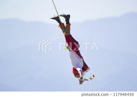 Mexican dance of the flyer, volador is hanging on the cable from the pole and performing the ancient ritual. Sea and mountains on the background. Mexican dance of the flyer, volador is hanging on the cable from the pole and performing the ancient ritual. Sea and mountains on the background. 117946430