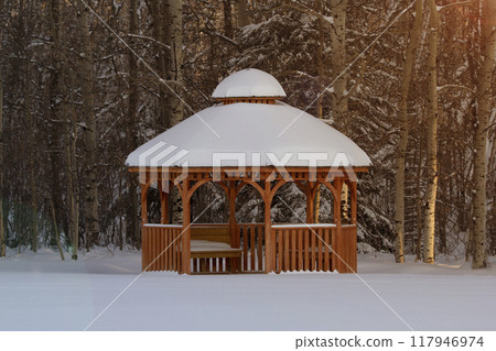 Rustic scene of the wooden cedar brown gazebo in the winter forest covered with snow, warm sunset light. Rustic scene of the wooden cedar brown gazebo in the winter forest covered with snow, warm sunset light. 117946974