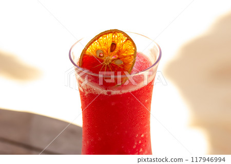 Bright red strawberry cocktail with crushed ice and dry orange slice on the wooden table on the beach of the resort, sunny day. 117946994