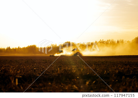 Tractor with a seeder is working in the dust in golden sunset light. 117947155