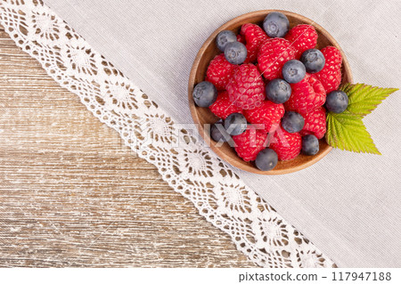 Plate with raspberries and blueberries on the wooden table with laced cloth. 117947188