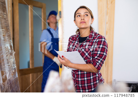 Young Latin woman in plaid shirt making notes on the plan of an apartment during renovation 117948785