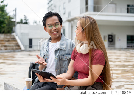 Two cheerful Asian college students are having a good conversation while relaxing outdoors on campus Two cheerful Asian college students are having a good conversation while relaxing outdoors on campus 117948849
