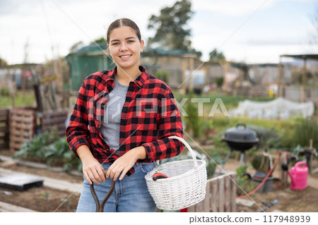 Smiling young female farmer posing with basket and garden instruments during season of sowing on sunny day of autumn Smiling young female farmer posing with basket and garden instruments during season of sowing on sunny day of autumn 117948939