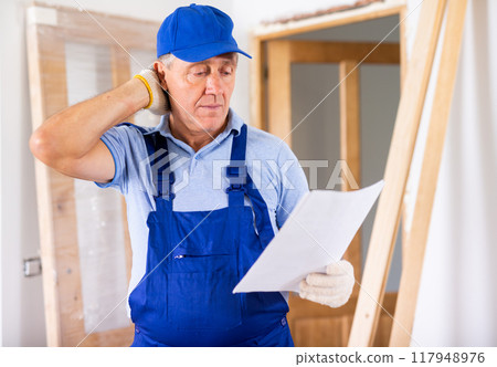 Serious middle-aged man in uniform exploring a plan of an apartment during renovation Serious middle-aged man in uniform exploring a plan of an apartment during renovation 117948976
