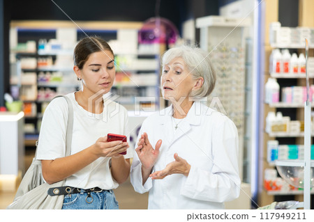 Senior female pharmacist communicates with girl client, looks at mobile phone screen Senior female pharmacist communicates with girl client, looks at mobile phone screen 117949211