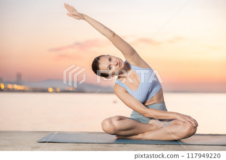 Near sea during sunset, girl performs variations of padmasana with body tilt 117949220