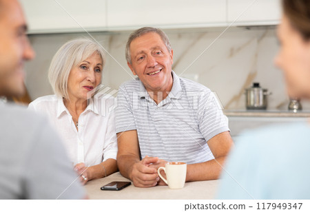 Smiling parents and adult children sit at the table with tea and talk about home life Smiling parents and adult children sit at the table with tea and talk about home life 117949347