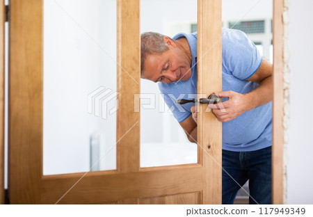Ordinary elderly man independently installs bronze door handles on wooden door 117949349