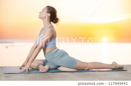 Near sea during sunset, girl performs variations of pigeon pose 117949441