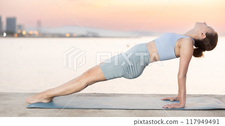Near sea during sunset, girl performs reverse plank pose Near sea during sunset, girl performs reverse plank pose 117949491