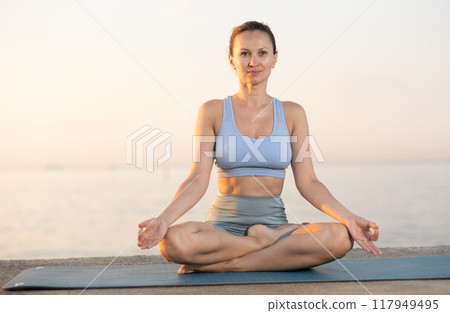 Happy slim woman making yoga meditation in lotus pose on beach at sunrise 117949495