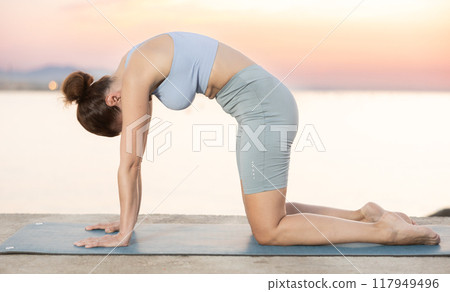 Portrait of caucasian woman standing on sports mat and making yoga meditation in cat-cow pose of yoga on beach at morning 117949496