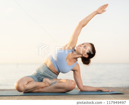 Happy young experienced woman fitness teacher performs yoga parshvottanasana sitting on beach at morning Happy young experienced woman fitness teacher performs yoga parshvottanasana sitting on beach at morning 117949497