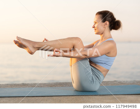 Diligent young woman trainer practicing upward boat pose of yoga, ubhai padangushthasan on mat on seashore at sunset. Philosophy of active lifestyle Diligent young woman trainer practicing upward boat pose of yoga, ubhai padangushthasan on mat on seashore at sunset. Philosophy of active lifestyle 117949498
