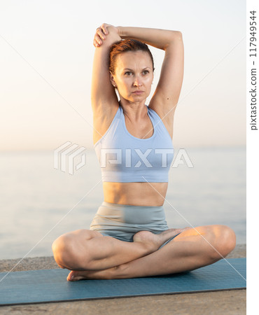 Portrait of caucasian woman sitting on sports mat and making yoga meditation in lotus pose and hands up on seashore at sunrise 117949544