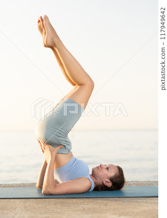 Experienced sporty young woman teacher performs feathered peacock asana, pincha mayurasana on beach at sunrise 117949642