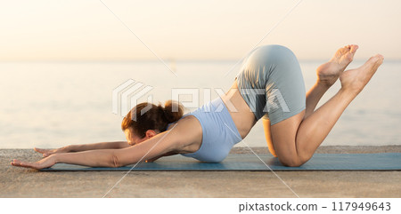 Skilled young female trainer performing cat-cow pose of yoga on seashore at sunrise 117949643