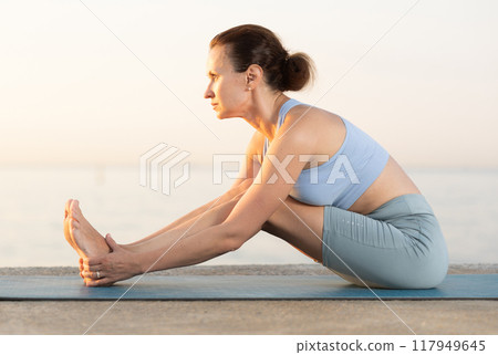 Sporty energetic young woman in sportwear training stretching legs workout on beach at sunrise 117949645