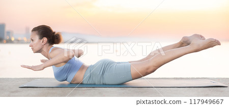 Near sea during sunset, girl performs shalabhasana 117949667